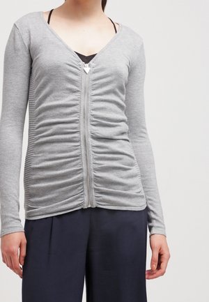 Vrouw draagt een gerimpeld grijs zip-up top met lange mouwen over een zwarte bralette, gecombineerd met losse zwarte broek tegen een effen achtergrond.