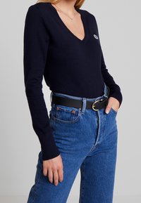 Navy V-neck sweater with long sleeves, worn with high-waisted blue jeans and a black belt featuring a gold buckle. Smooth texture, fitted design.