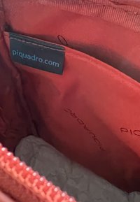 Interno in tessuto rosso di una borsa con etichetta nera riportante "piquadro.com." Include una piccola tasca e carta accartocciata sul fondo.
