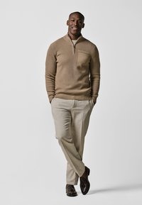 Brauner gerippter Strickpullover mit Reißverschlusskragen und Tasche, kombiniert mit hellbeigen Hosen und dunklen Loafers. Schlichte und moderne Stilrichtung.