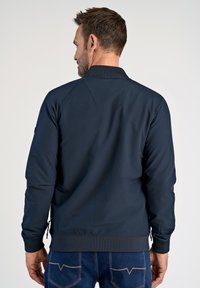 Granatowa kurtka bombowca z prążkowanym kołnierzem i mankietami, gładkim materiałem oraz zamkniętą kieszenią na lewym rękawie.