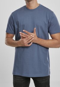 Muž v modrém tričku, který tlačí svou pravou ruku levou rukou před jednobarevným šedým pozadím.