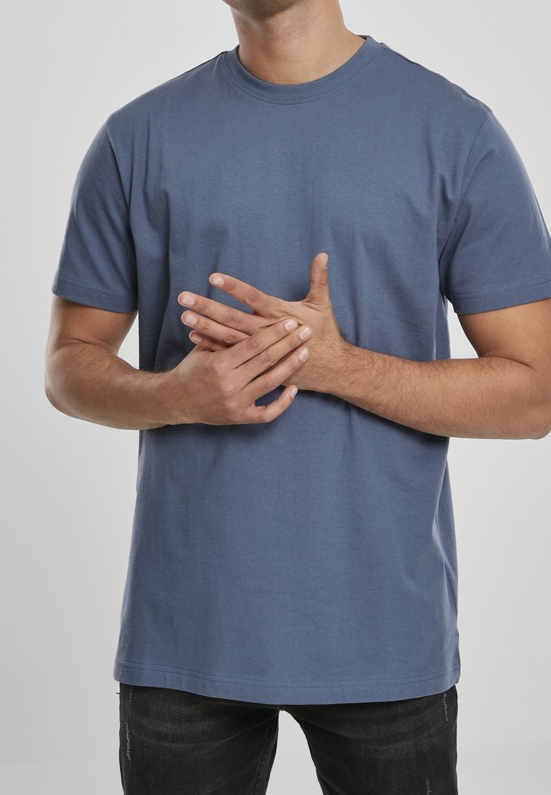 Muž v modrém tričku, který tlačí svou pravou ruku levou rukou před jednobarevným šedým pozadím.
