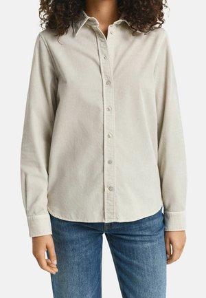 Kobieta ubrana w beżową koszulę ze sztruksu zapinaną na guziki z długim rękawem i niebieskie dżinsy, stojąca na jednolitym tle.