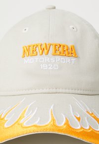 Beige keps med broderad "NEW ERA" i orange, "MOTORSPORT" i vitt, samt flamtmönster i orange och vitt längs skärmen.