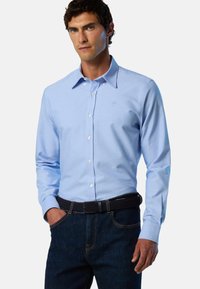 Camicia azzurro chiaro in cotone con maniche lunghe, colletto appuntito e piccolo logo sul petto a sinistra. Indossata con jeans scuri.