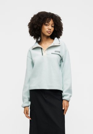 Femme aux cheveux bouclés portant un pull en polaire bleu clair avec une poche poitrine zippée et une jupe noire sur un fond uni.