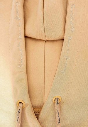 Beige hoodie van een textuurstof. Voorzien van een trekkoordsluiting met houten uiteinden en geborduurde accenten langs de randen.