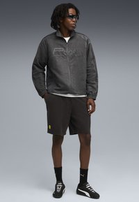 Sort ruskinds zip-jakke med hævet Puma-logo, parret med mørke shorts med et gult Ferrari-emblem, suppleret med sorte sneakers.