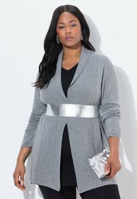 Grå stickad cardigan med långa ärmar, öppen front och ett metalliskt silverfärgat midjebälte. Modellen håller en liten silverfärgad kuvertväska.