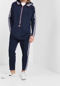 Personen bär en marinblå Nike träningsoverall med logotape på ärmarna och byxornas sidor, i kombination med vita Nike sneakers.