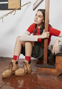 Botas de tobillo de ante marrón con suelas gruesas y texturizadas y cordones burdeos. El modelo lleva calcetines rojos y una blusa roja de canalé, sentada contra una estructura de madera.