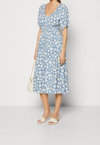 Vrouw in een blauwe midi-jurk met witte bloemenprint, puffmouwen, gerimpelde taille, die een witte handtas vasthoudt en crèmekleurige hakken draagt.