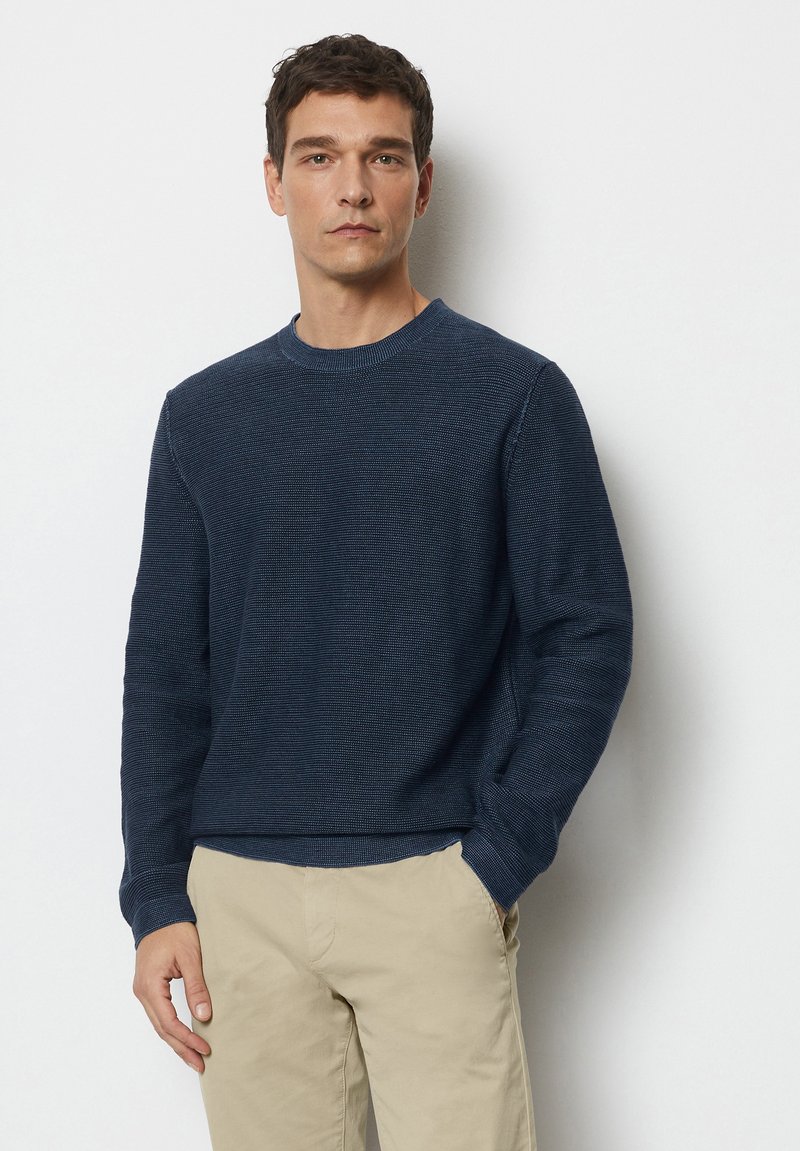 Marineblauer Rundhalspullover mit strukturiertem Strick, langen Ärmeln und gerippten Bündchen; kombiniert mit beigen Hosen für einen lässigen Look.
