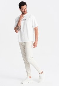 Camiseta blanca de algodón con texto gráfico negro, combinada con pantalones de chándal en color crema que tienen una textura suave y puños acanalados, y zapatillas blancas.