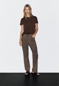 Brun kortærmet t-shirt parret med brune suede lige bukser og sorte åbne sandaler, placeret mod en enkel hvid baggrund.