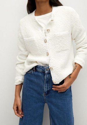 Kvinde iført en hvid, tekstureret cardigan med perleagtige knapper, hvid skjorte under og blå højttaljede jeans.