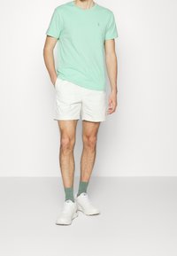 Muntgroene katoenen T-shirt met een blauw logo, gecombineerd met witte shorts, groene sokken en witte sneakers. Eenvoudig ontwerp zonder patronen.