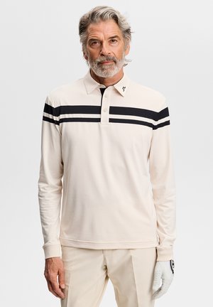 Uomo di mezza età con capelli e barba grigi, che indossa una polo beige a maniche lunghe con strisce nere e un guanto da golf bianco sulla mano sinistra.