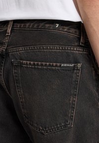 Jeans in denim nero con una texture scolorita, dotati di un'etichetta sulla tasca posteriore e di un'etichetta numerata sul girovita. Cuciture in colori a contrasto.