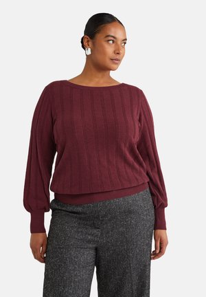 Maglione lavorato a maglia bordeaux con costine verticali, scollatura ampia e maniche a sbuffo. Abbinato a pantaloni grigi scuri con texture.