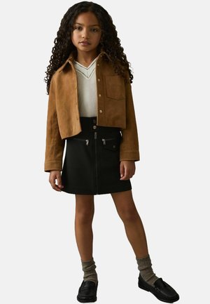 Giacca in suede marrone corta sopra un maglione bianco, abbinata a una gonna nera con zip e scarpe nere con calzini grigi.