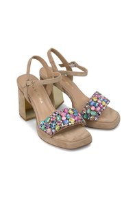 Beige suède hoge hak sandalen hebben een brede versierde band versierd met levendige, multikleurige edelstenen en een goudkleurige blokhak.