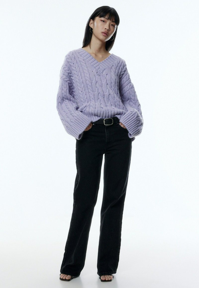 Lavendelfarbenes Grobstrickpullover mit V-Ausschnitt und weiten Ärmeln, kombiniert mit schwarzen, geraden Jeans und schwarzen Sandalen mit offenem Zeh.