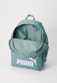 Tamsiai mėlyna kuprinė su sulankstomu viršumi, didele pagrindine kišenė, kvėpuojanti tinklinio šalutinė kišenė ir balta Puma logotipas priekyje.
