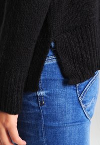 Mustat neulepaidat, joissa on teksturoitu pinta ja ribbireuna, osittain työnnettyinä sinisiin farkkuihin. Sivureunan yksityiskohta näkyy.