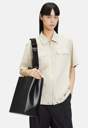Frau mit glattem schwarzen Haar trägt beige kurzärmeliges Hemd mit Knöpfen, schwarze Hose und trägt eine große schwarze Ledertasche mit breitem Trageriemen.