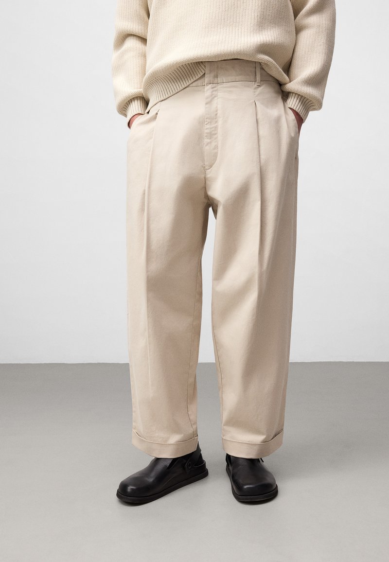 Beige cotton trousers with a loose fit and single pleat in front, paired with black slip-on shoes. Textured knit sweater visible.
