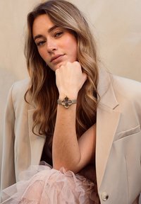 Bracelet en cuir gris avec éléments en métal doré, présentant un accent en forme de fleur au centre. La mannequin porte un blazer beige et une jupe en tulle.