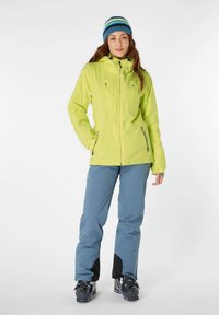 Heldere gele waterdichte jas met capuchon, met ritszakken; gecombineerd met lichtblauwe skibroek, zwarte accenten en zwarte snowboardboots.