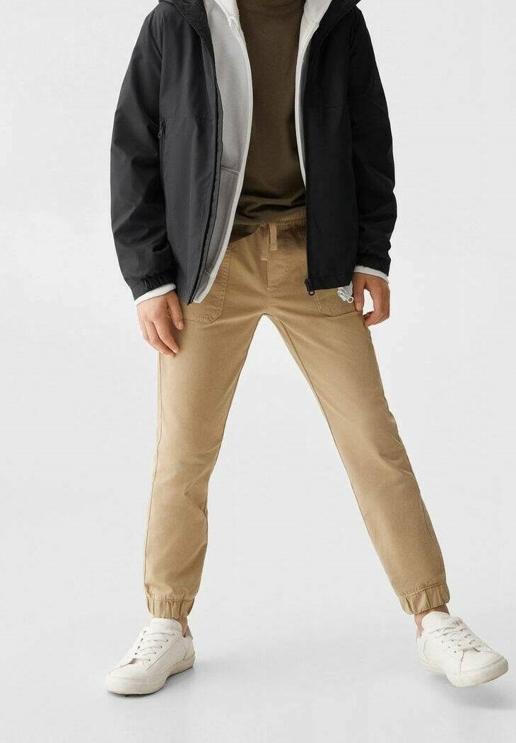 Svart zip-up-jacka över en grå tröja, ihop med beige avsmalnande byxor och vita sneakers. Slät tyg, elastiska muddar på byxorna.