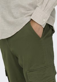 Olivgrüne Cargo-Shorts mit Seitentaschen, kontrastierender Naht und umgeschlagenem Saum. Kombiniert mit einem hellbeigen Langarmshirt.
