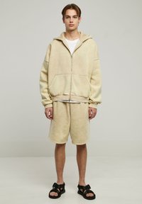 Beige cotton zip-up hoodie with front pockets, paired with matching shorts. Model wears black sandals. Plain background, neutral colors.