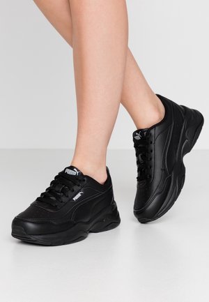 Person iført sorte Puma-sneakers med tykke såler, stående på en lysegrå gulv mod en ensfarvet lys baggrund.