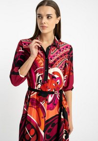 Kobieta ubrana w kolorową, wzorzystą sukienkę w odcieniach czerwieni, pomarańczu, różu i czerni, z zapięciem przy dekolcie i czarnym paskiem w talii, patrzy na bok.