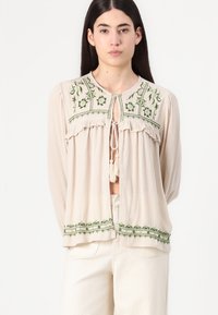 Blouse beige en tissu texturé, broderie florale verte au niveau du col et de l'ourlet, et fermeture à lien frontal avec des pampilles. Manches longues incluses.