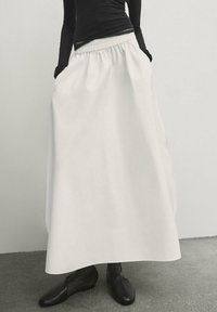 Gonna bianca lunga fino alla caviglia con vita elastica e tasche laterali, realizzata in tessuto leggero. Abbinata a scarpe nere a pianta piatta.