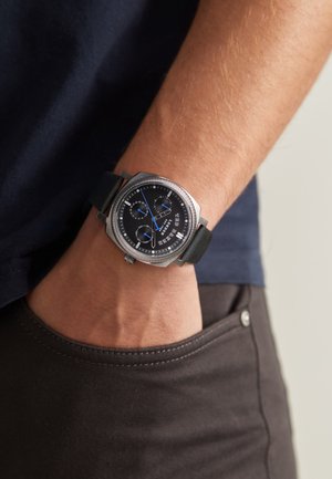 Montre chronographe argentée avec un bracelet en cuir noir, cadran rond, accents bleus et lunette texturée, portée au poignet.