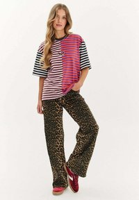 Kobieta z długimi falowanymi włosami, ubrana w pół różową/pół czarną oraz pół pomarańczową/pół purpurową w paski koszulę z naszywką w kształcie motyla, spodnie w cętkowany wzór oraz czerwone trampki.