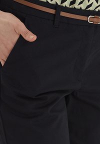 Pantalon noir en tissu lisse, avec des poches latérales et une ceinture en cuir marron avec une boucle en argent, main positionnée dans la poche.