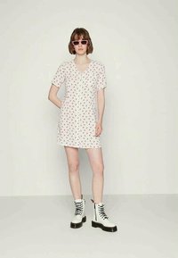 Mulher vestindo um vestido curto branco com estampado floral vermelho, botas brancas com atacadores e óculos de sol cor-de-rosa, em pé contra um fundo simples.