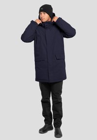 Parka isolata blu navy con colletto alto, doppie tasche anteriori, chiusura con zip e cappuccio regolabile, abbinata a pantaloni neri e stivali.