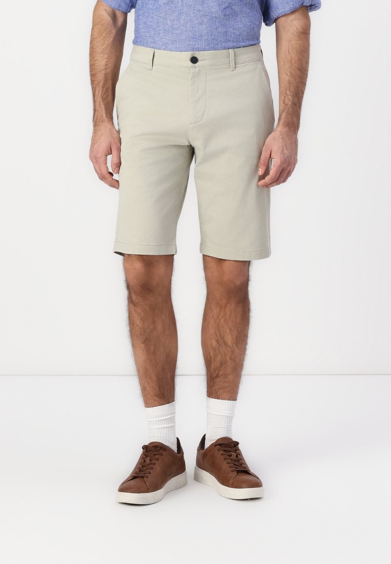 Homme portant un short beige jusqu'au genou, des chaussettes blanches, des baskets basses marron et une chemise bleue à manches courtes, debout devant un fond blanc.