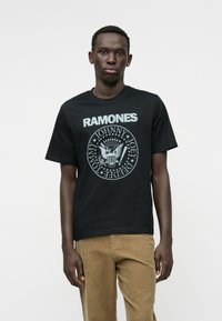 Melna kokvilnas T-kreklā ir uzraksts "RAMONES" baltā krās�ā krūšu daļā un apaļa grafika ar ērgli un nosaukumiem gaiši pelēkā krāsā.
