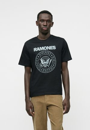 Zwarte katoenen t-shirt met de tekst "RAMONES" in witte letters op de borst en een ronde grafiek met een adelaar en namen in lichtgrijs.