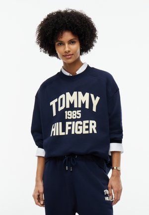 Giovane donna con capelli ricci che indossa una felpa Tommy Hilfiger blu navy e pantaloni abbinati, in piedi contro uno sfondo bianco semplice.